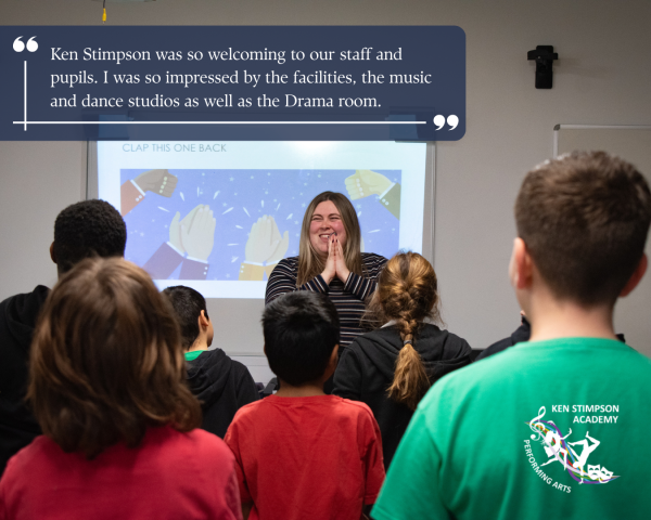 Photo of a teacher stood before a class clapping and smiling. The students have their backs to the camera