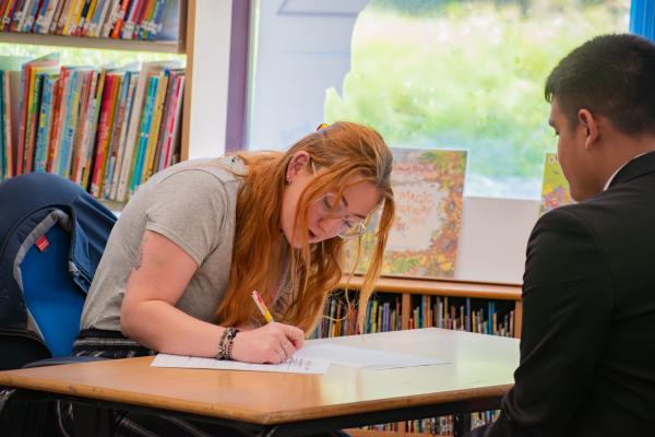 Photo of a student and a business volunteer sat at a desk.  The volunteer is writing something on a sheet of a paper.  The student has their back to the camera.  They are in a library next to a window with a bookcase behind them.