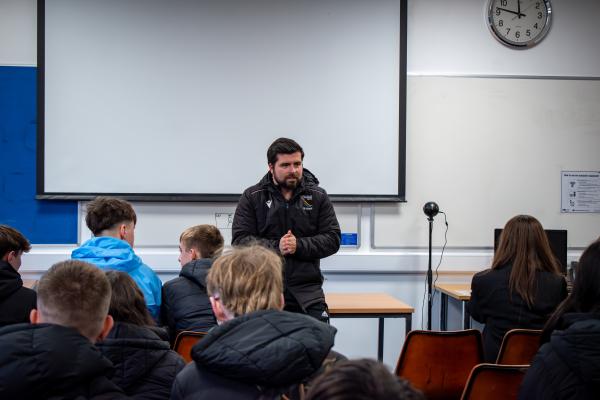 Photo of a group of students sat in rows watching as Mr Isaacs addresses them at the front of the room.