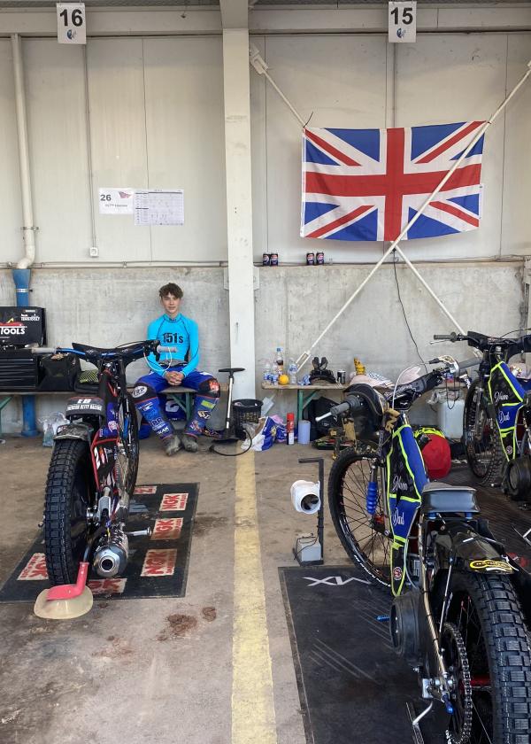 Photo of a speedway rider sat at the back of a garage with two bikes in front of him